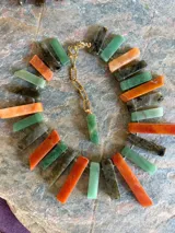 Collar corto elaborado con piedras rectangulares en tonos verdes, naranjas y grises oscuros, unidas con herrajes dorados y cadena de eslabones con dije colgante.