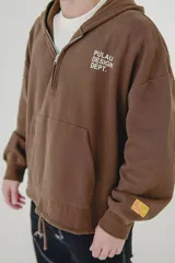 Canguro beige con capucha, cierre frontal parcial y bolsillo tipo canguro. Presenta estampado con texto en el pecho y detalle en la manga.