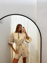 Mono corto de lino color beige, con cuello camisero, cierre frontal, mangas largas abullonadas y cinto ajustable en la cintura.