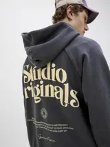 Canguro negro con capucha, de corte holgado y mangas raglán. Presenta un gran estampado en color beige en la espalda con la inscripción "Studio Originals" y texto descriptivo, además de puños y dobladillo acanalados.