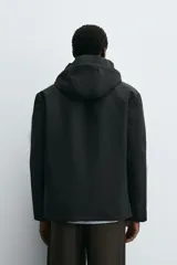 Cazadora técnica regular fit de color negro, con interior ligeramente acolchado. Presenta cuello subido con capucha ajustable, cierre frontal de cremallera oculta por solapa con botones a presión, bolsillos laterales y puños con interior de rib.