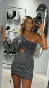 Vestido corto ajustado de una manga larga, color negro con brillo plateado. Presenta un escote asimétrico de un hombro y un corte tipo cut-out en la zona de la cintura.