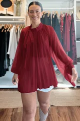 Blusa plisada de corte amplio y fluido, color bordó, con cuello alto y lazo decorativo. Presenta mangas largas con volumen y puños ajustados.