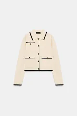 Chaqueta de punto color crema con ribetes en contraste negro, cuello solapa, manga larga y cierre frontal con botones dorados. Presenta bolsillos de plastrón en el delantero con detalle de botones.