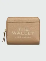 Billetera compacta Marc Jacobs modelo 'The Leather Mini Compact Wallet', color beige claro, con textura de cuero granulado y cierre de cremallera. Presenta el texto 'THE WALLET MARC JACOBS' grabado en el frente.