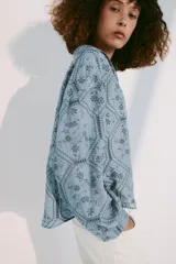 Camisa azul con cuello de solapa, escote en pico y mangas largas con puños abotonados. Presenta un diseño con bordados perforados y patrones geométricos y florales. Cierre frontal con botones.