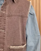 Chaleco de denim color marrón, confeccionado en 100% algodón, con bolsillos frontales amplios, cierre de botones y costuras decorativas bordadas en blanco en los bordes y los bolsillos.