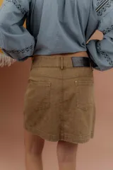 Minifalda de gabardina color tostado, de tiro medio con bolsillos delanteros y traseros, presillas para cinto y cierre frontal con botón metálico.