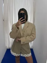 Conjunto de dos piezas color beige compuesto por un blazer corto con apliques metálicos tipo alfileres y un short con detalle de puntilla blanca en el ruedo.