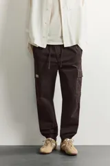 Pantalón cargo de corte ajustado en color negro, confeccionado en tejido de algodón con elasticidad. Presenta cintura elástica con cordón ajustable, bolsillos laterales tipo cargo con solapa y bajo con puños elásticos.