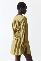 Vestido corto oversized de mezcla de viscosa y color verde militar, con escote en V profundo, cordón ajustable en el cuello y mangas globo largas con puños elásticos. Presenta fruncidos en el canesú y sección de punto smock en la cintura con falda de leve vuelo.