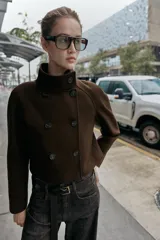 Chaqueta corta de paño en color beige, con cuello subido, manga larga y cierre frontal cruzado con botones oscuros. Presenta bolsillos delanteros de vivo.