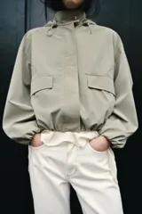 Cazadora de cuello subido con capucha ajustable, manga larga con puños elásticos y bolsillos delanteros con solapa. Presenta bajo elástico y cierre frontal con cremallera oculta y botones a presión.