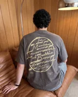 Remera de corte oversize color marrón oscuro, con estampa gráfica circular en color crema en la espalda que presenta tipografía estilizada y detalles de texto en la parte inferior.