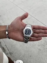 Reloj de pulsera con diseño inspirado en el modelo Nautilus, con caja y malla de acero inoxidable. Presenta una esfera azul con textura estriada horizontal, índices aplicados luminiscentes y ventana de fecha a las tres. Incluye cierre desplegable tipo mariposa.