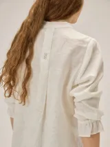 Camisa de lino color crudo, con cuello mao y cierre frontal con botones. Cuenta con detalles de alforzas verticales y diseño bordado floral en el frente, además de mangas 3/4 con volumen y puños elásticos.