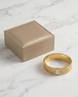 Esclava rígida de acero con baño de oro, con diseño de malla hexagonal calada. Presenta un aplique central rectangular con incrustaciones de circonias y centro de nácar.