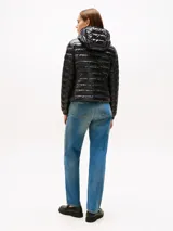 Campera puffer ultraliviana de color negro con acabado brillante, diseño acolchado horizontal y capucha. Presenta cierre frontal de cremallera, bolsillos laterales con cierre y logo engomado de Tommy Hilfiger en la manga. Confeccionada en nailon reciclado con corte regular.