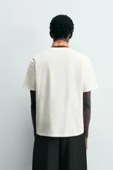 Camiseta de corte relajado (relaxed fit) color blanco hueso, con cuello redondo, manga corta y bolsillo plastrón en el pecho. Presenta un bordado pequeño y contrastante de una palmera en el área del bolsillo.