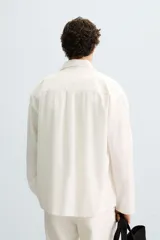 Camisa de corte relajado, color blanco hueso, con finas rayas verticales en tono grisáceo. Presenta cuello solapa, manga larga, bolsillo de parche en el pecho y bajo con aberturas laterales.