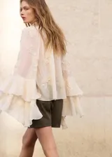 Blusa color marfil de tejido fluido, con cuello alto con lazo, volados frontales y mangas largas acampanadas con detalles florales bordados.