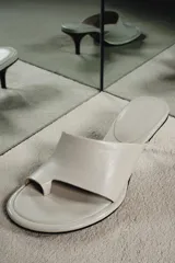 Sandalia tipo mule de tacón alto fino de madera, confeccionada en piel color blanco marfil. Presenta una tira asimétrica con separador para el dedo y punta redonda.