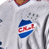 Camiseta de fútbol Umbro del Club Nacional de Football, modelo Home 2026. Diseño en color blanco con tejido jacquard que presenta un patrón de trofeos históricos en toda la prenda. Incluye cuello con detalles tricolores, logo de Umbro, escudo del club con estrellas y sponsors estampados en el frente y mangas.