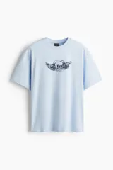 Remera celeste de corte holgado, con cuello redondo y diseño gráfico estampado en el pecho que incluye alas y texto.