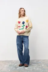 Sweater de punto color crudo con diseño de flores y formas geométricas bordadas en colores vibrantes. Presenta cuello redondo, mangas largas con detalles de líneas laterales en contraste y corte holgado.