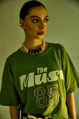 Remera verde con estampado frontal con la frase "The Muse" y el número 85 en blanco y lila. De corte holgado y largo a la cadera.