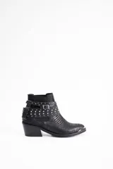 Botín de cuero vacuno negro con textura grabada, taco cuadrado y punta fina con detalle metálico. Presenta una tira decorativa con tachas metálicas y hebilla ajustable alrededor del tobillo.
