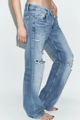 Jeans de tiro bajo y cinco bolsillos, con diseño de pierna ancha y larga. Presenta roturas y desgastes sutiles en la parte delantera. Cierre frontal con cremallera y botón metálico.