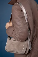 Cartera de hombro color bordó, confeccionada en cuero con acabado brillante. Presenta detalles de remaches metálicos plateados en los laterales y la base, y una correa ajustable con hebilla. Incluye una placa metálica con el logo de la marca en la parte frontal.