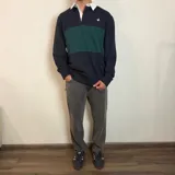 Buzo tipo polo de rugby con cuello blanco, manga larga y puños acanalados. Presenta un diseño de bloques de color en azul marino y verde, con un pequeño logo bordado en el pecho.