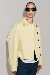 Chaqueta corta confeccionada en mezcla de algodón color amarillo claro, con cuello de solapa, manga larga, bolsillos delanteros y cierre frontal con botones visibles en los extremos.