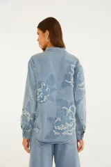 Sobrecamisa de denim azul con intrincados bordados florales en relieve, cierre frontal con botones metálicos y bolsillos delanteros con solapa.