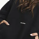 Buzo tipo hoodie color marrón, con capucha, bolsillo frontal tipo canguro y estampado minimalista en el pecho. Presenta un diseño holgado y mangas largas.