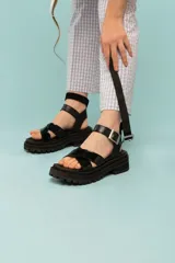 Sandalias negras de estilo chunky con tiras cruzadas en charol y base de goma dentada. Cuentan con una tira superior desmontable y cierre con hebilla metálica en el tobillo.