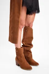 Bota de caña alta confeccionada en cuero vacuno, con diseño versátil que permite doblar la caña. Presenta un taco cuadrado de altura media y punta redondeada.