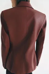 Blazer de cuero sintético en color beige, con cuello solapa, manga larga y bolsillos con solapa en el frente. Cierre frontal cruzado con botones a tono.