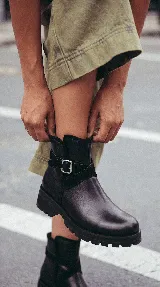Bota corta de cuero negro con suela track de goma, detalle de tira cruzada con hebilla metálica lateral y paneles de gamuza en contraste.