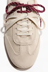 Calzado deportivo de gamuza color beige, con cordones intercambiables en tonos blanco y bordó, detalle de talón plegable con refuerzo negro y suela plana de goma acanalada.