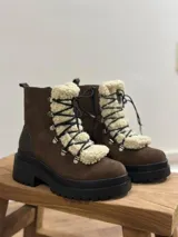Bota tipo borcego de cuero marrón con suela track negra de goma. Presenta un aplique frontal desmontable de corderito sintético color crema y cordones negros con ganchos metálicos.
