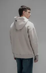 Hoodie oversize de color beige, confeccionado en frisa premium. Presenta capucha ajustable con cordones, bolsillo tipo canguro frontal y puños con aberturas para pulgares.