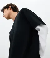 Remera de corte oversized con diseño de doble capa, que combina una remera negra de manga corta con mangas largas blancas integradas debajo. Confeccionada en algodón, presenta un estilo urbano y moderno.