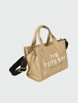 Bolso tipo tote de lona color beige de la marca Marc Jacobs. Presenta el texto 'THE TOTE BAG' estampado en el frente en color blanco, dos asas de mano superiores y una correa de hombro ajustable y removible en color negro.