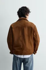 Cazadora de piel con acabado ante color marrón, corte relaxed fit, cuello bowling y manga larga con puño abotonado. Presenta bolsillos de vivo en la cadera y cierre frontal de botonadura.