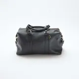 Bolso de mano estilo bowling en color negro, confeccionado en material texturizado. Presenta doble asa superior con detalles de hebillas decorativas, cierre principal con cremallera y correa larga ajustable y extraíble para llevar al hombro o cruzado. Incluye herrajes metálicos en tono dorado.
