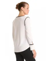 Blusa de crepe negro con ribetes en color beige que recorren el escote, la línea central y las costuras de los hombros. Presenta cuello redondo con abertura en V, mangas largas con puños abiertos y detalle de botón metálico, y ruedo redondeado.