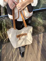 Bolso tipo tote color crema, con detalles en cuero color marrón. Tiene un bolsillo con cierre en la parte superior y un parche de cuero con el logo de la marca en la parte inferior.